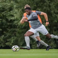 Joven futbolista practica ejercicios de conduccion de balon en el campo, con un maniquí de entrenamiento y otro jugador detrás.
