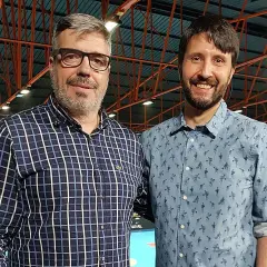 Dos hombres sonríen a la cámara. El de la izquierda, con gafas y camisa a cuadros, es Paco Caro. El de la derecha lleva una camisa azul con un estampado divertido.