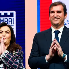 Nita Ambani y un hombre sonríen con las manos juntas, con el logo de la Superliga India de fondo.