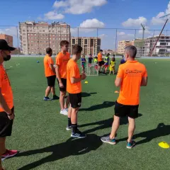 Entrenadores con camisetas naranjas dirigen a jóvenes deportistas en un campo de fútbol. Uno de ellos, con gorra negra, parece estar explicando algo, quizás sobre la equivalencia de técnico deportivo nivel 1.