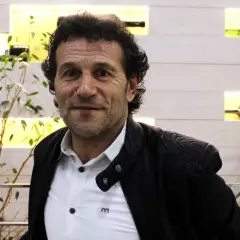 Luisito entrenador, un hombre de mirada intensa, posa con una chaqueta negra sobre una camisa blanca. Detrás, estanterías iluminadas con botellas y plantas.