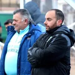 Dos entrenadores, uno mayor con pelo gris y otro más joven con barba, observan el partido. El más joven, con los brazos cruzados, parece concentrado. El mayor, con una chaqueta azul, viste una camiseta clara con texto. Ambos parecen estar en un evento ...