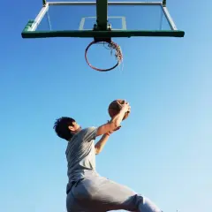 Joven se prepara para la entrada a canasta de baloncesto, con el balón en alto contra un cielo azul.