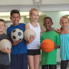 Cinco niños sonríen, cada uno con un balón. Uno sostiene un balón de baloncesto, listo para jugar.