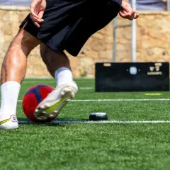 Jugador de fútbol entrenando ejercicios de velocidad en campo artificial, con balón y dispositivo de medición.