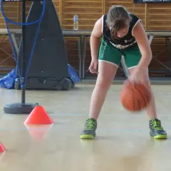 Niña concentrada botando el balón de baloncesto entre conos naranjas en un gimnasio.