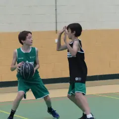 Dos niños juegan al baloncesto. Uno, con camiseta verde, se prepara para hacer un pase de pecho. El otro, con camiseta negra, se defiende.