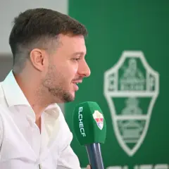 El entrenador del Elche CF, con camisa blanca, habla a un micrófono verde con el escudo del club.