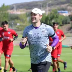 Entrenador sonriente lidera a jugadores en un partido pretemporada. Corren en un campo verde bajo el sol.