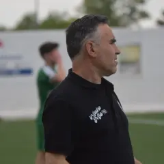 Entrenador de fútbol con polo negro, mirando hacia la derecha, con un jugador de verde al fondo.
