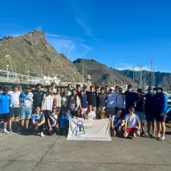 Grupo de jóvenes y adultos posan con una bandera del **CD Urda** en un puerto deportivo, con montañas y barcos de fondo.