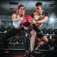 Pareja realizando ejercicios en pareja con pelota en un gimnasio, rodeados de pesas.