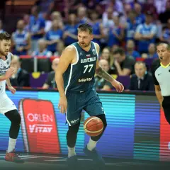 Jugador esloveno con el número 77 dribla el balón, mientras un árbitro observa atentamente. El jugador islandés, con el número 7, se prepara para defender. Se percibe una falta en ataque en el baloncesto.