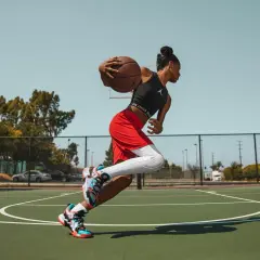 Jugadora de baloncesto en movimiento, con el balón en las manos, lista para anotar. Sus aleros de baloncesto brillan bajo el sol.