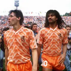 Dos futbolistas de equipos holandeses con camisetas naranjas icónicas, rodeados de aficionados y cámaras.