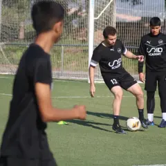 Entrenador enseña a joven cómo mejorar en fútbol, practicando pases en campo.