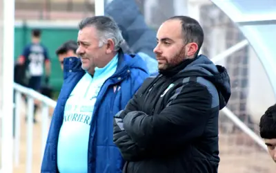 Dos entrenadores, uno mayor con pelo gris y otro más joven con barba, observan el partido. El más joven, con los brazos cruzados, parece concentrado. El mayor, con una chaqueta azul, viste una camiseta clara con texto. Ambos parecen estar en un evento ...