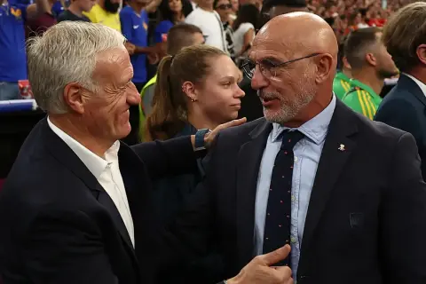 Dos entrenadores, uno con traje oscuro y corbata, el otro con traje azul, se saludan. Podría ser un momento de tensión o respeto mutuo tras un partido entre España y Francia.
