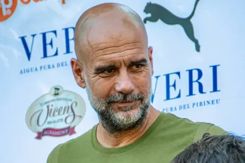 Pep Guardiola, con su mirada reflexiva, parece estar en un evento de "pep guardiola estudios". Detrás, logos de "Veri" y "Vicens".