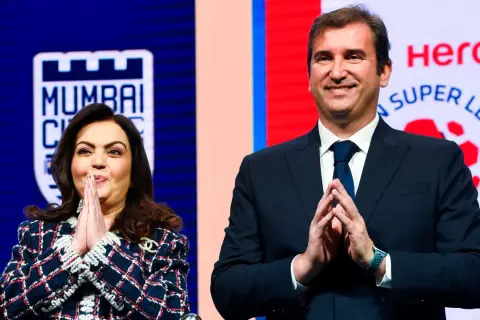Nita Ambani y un hombre sonríen con las manos juntas, con el logo de la Superliga India de fondo.