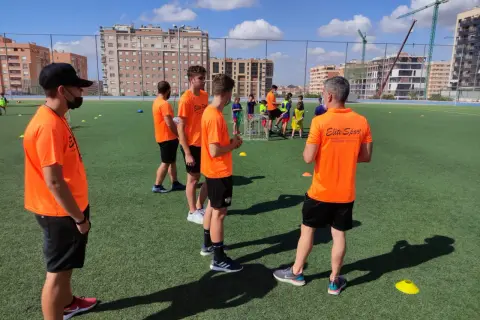 Entrenadores con camisetas naranjas dirigen a jóvenes deportistas en un campo de fútbol. Uno de ellos, con gorra negra, parece estar explicando algo, quizás sobre la equivalencia de técnico deportivo nivel 1.