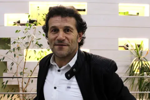 Luisito entrenador, un hombre de mirada intensa, posa con una chaqueta negra sobre una camisa blanca. Detrás, estanterías iluminadas con botellas y plantas.