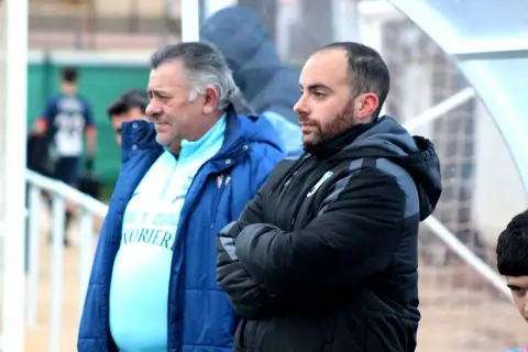 Dos entrenadores, uno mayor con pelo gris y otro más joven con barba, observan el partido. El más joven, con los brazos cruzados, parece concentrado. El mayor, con una chaqueta azul, viste una camiseta clara con texto. Ambos parecen estar en un evento ...