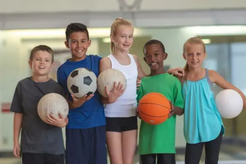 Cinco niños sonríen, cada uno con un balón. Uno sostiene un balón de baloncesto, listo para jugar.
