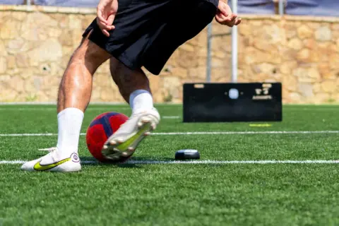 Jugador de fútbol entrenando ejercicios de velocidad en campo artificial, con balón y dispositivo de medición.