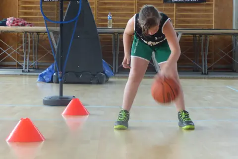 Niña concentrada botando el balón de baloncesto entre conos naranjas en un gimnasio.