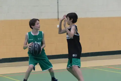 Dos niños juegan al baloncesto. Uno, con camiseta verde, se prepara para hacer un pase de pecho. El otro, con camiseta negra, se defiende.