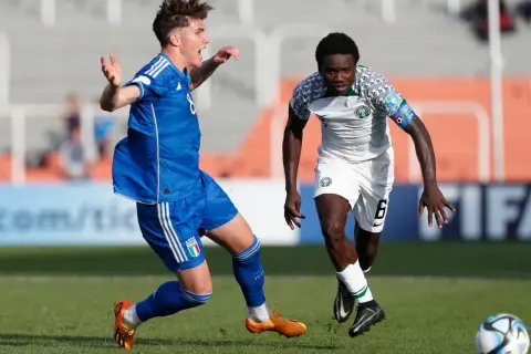 Jugadores de fútbol en acción, uno con camiseta blanca y verde de la **selección Nigeria**, el otro con azul.