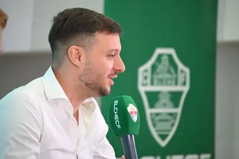 El entrenador del Elche CF, con camisa blanca, habla a un micrófono verde con el escudo del club.