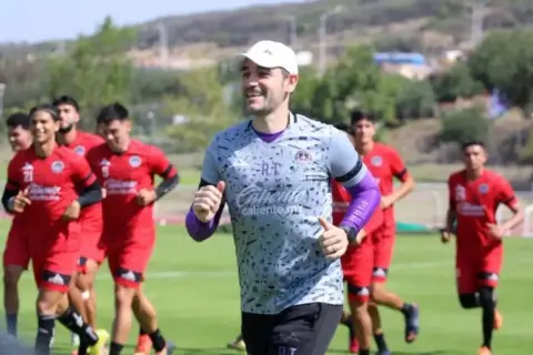 Entrenador sonriente lidera a jugadores en un partido pretemporada. Corren en un campo verde bajo el sol.