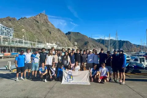 Grupo de jóvenes y adultos posan con una bandera del **CD Urda** en un puerto deportivo, con montañas y barcos de fondo.