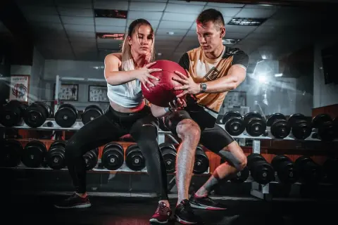 Pareja realizando ejercicios en pareja con pelota en un gimnasio, rodeados de pesas.