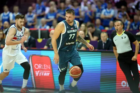 Jugador esloveno con el número 77 dribla el balón, mientras un árbitro observa atentamente. El jugador islandés, con el número 7, se prepara para defender. Se percibe una falta en ataque en el baloncesto.