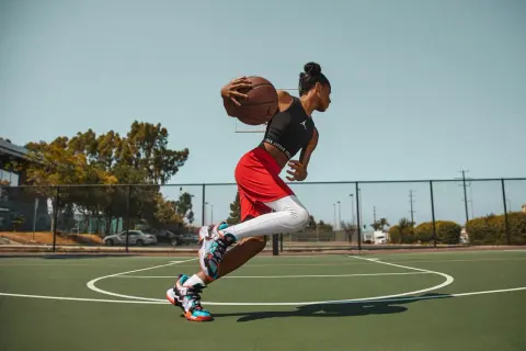 Jugadora de baloncesto en movimiento, con el balón en las manos, lista para anotar. Sus aleros de baloncesto brillan bajo el sol.