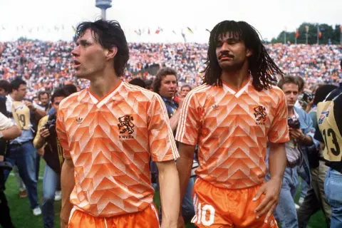 Dos futbolistas de equipos holandeses con camisetas naranjas icónicas, rodeados de aficionados y cámaras.
