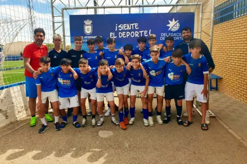 Equipo de fútbol infantil posa sonriente. El cartel de fondo anuncia "¡Siente el deporte!" y el logo del Ayuntamiento de San José.