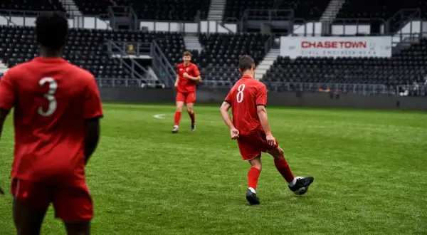 Jugadores de fútbol en uniforme rojo practicando ejercicios de pases en un campo verde, con gradas vacías al fondo.