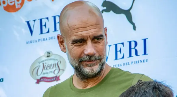 Pep Guardiola, con su mirada reflexiva, parece estar en un evento de "pep guardiola estudios". Detrás, logos de "Veri" y "Vicens".
