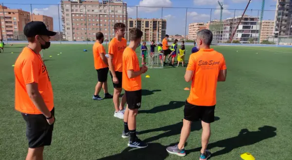 Entrenadores con camisetas naranjas dirigen a jóvenes deportistas en un campo de fútbol. Uno de ellos, con gorra negra, parece estar explicando algo, quizás sobre la equivalencia de técnico deportivo nivel 1.