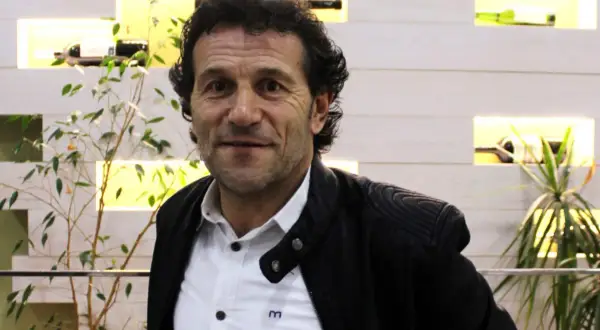 Luisito entrenador, un hombre de mirada intensa, posa con una chaqueta negra sobre una camisa blanca. Detrás, estanterías iluminadas con botellas y plantas.