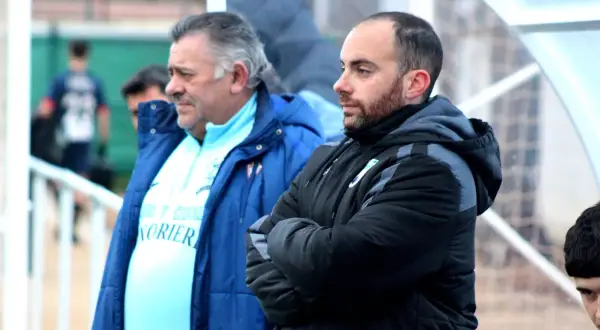 Dos entrenadores, uno mayor con pelo gris y otro más joven con barba, observan el partido. El más joven, con los brazos cruzados, parece concentrado. El mayor, con una chaqueta azul, viste una camiseta clara con texto. Ambos parecen estar en un evento ...