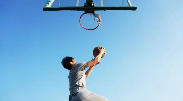 Joven se prepara para la entrada a canasta de baloncesto, con el balón en alto contra un cielo azul.