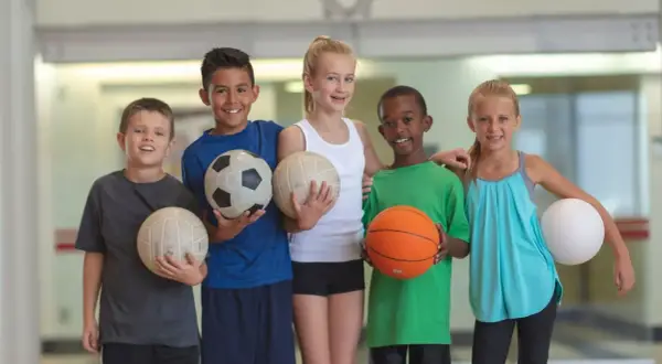 Cinco niños sonríen, cada uno con un balón. Uno sostiene un balón de baloncesto, listo para jugar.