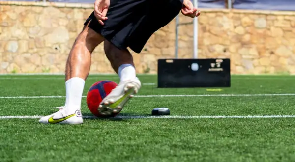 Jugador de fútbol entrenando ejercicios de velocidad en campo artificial, con balón y dispositivo de medición.