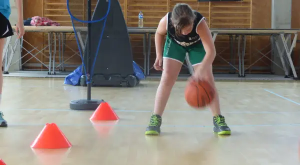 Niña concentrada botando el balón de baloncesto entre conos naranjas en un gimnasio.