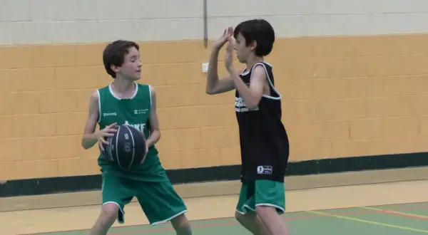 Dos niños juegan al baloncesto. Uno, con camiseta verde, se prepara para hacer un pase de pecho. El otro, con camiseta negra, se defiende.